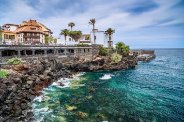 Tenerife 'deki Puerto de la Cruz' dan temiz, turkuaz suyu olan kayalık bir kıyı şeridi sahnesi. Palmiye ağaçlarıyla çevrili terasotta çatısı olan geleneksel bir bina denize bakan bir uçurumun kenarında oturur.
