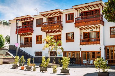 Los Gigantes, Tenerife 'de bulunan ahşap balkonlar ve pencere çerçeveleri de dahil olmak üzere geleneksel mimari özelliklere sahip üç katlı bir bina. Binanın önünde, saksı bitkileri ve ağaçlarla süslenmiş asfaltlı bir alan var.