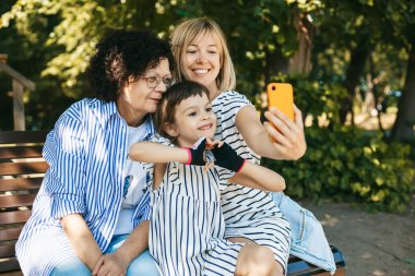 Aile zamanı: büyükanne, anne ve kız parktaki bankta selfie çekiyorlar, birlikte vakit geçiriyorlar.