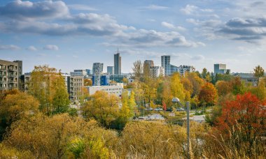 Vilnius, Litvanya 'nın sonbahardaki panoramik manzarası, ön planda canlı sarı ve portakal ağaçları, orta zeminde yerleşim ve ticari binaların bir karışımı ve açık bir gökyüzünün altındaki gökdelende egemen olan uzun boylu bir gökdelen.