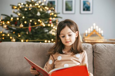 Genç bir kız, kanepede rahat rahat, büyük kırmızı bir kitap okuyor, arkasında altın ve kırmızı süslemelerle süslenmiş bir Noel ağacı duruyor.