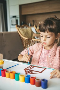 Küçük bir kız, elinde boya fırçası, bir masada oturuyor, birden fazla renge bürünmüş bir evi boyuyor, tamamen sanatına gömülmüş.