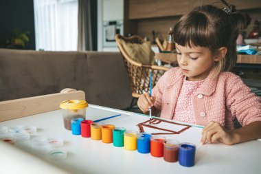 Masada resim yapan küçük bir çocuk. Gouache 'nin parlak renkleriyle çevrili. Yaratıcı projesine odaklanmış.