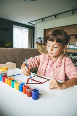 Masadaki küçük bir kız çok renkli guaj balığıyla resim yapıyor. Etrafı renkli boya kutularıyla çevrili. Sanat eserlerine odaklanıyor.