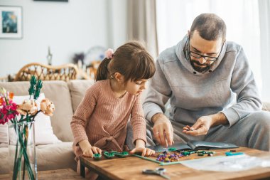 Oturma odasında 5-6 yaşlarında bir kız ve babası ahşap bir masada küçük inşaatlarla uğraşıyorlar.