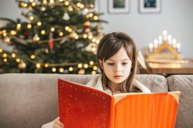 Kanepede kırmızı bir kitap okuyan küçük bir kızın Noel ağacı süslerle dolu sıcacık bir tatil anı.