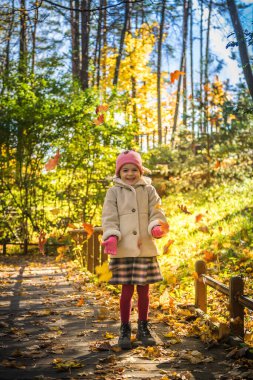 Bir sonbahar sahnesi. Ormanlık bir yolda duran gülen küçük bir kız. Etrafında renkli yapraklar düşerken sıcak güneş ışığıyla yıkanmış.