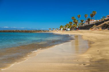 Tenerife 'den Playa Del Duque yumuşak kumlar ve parıldayan mavi sularla sakin bir sahne sunuyor. Berrak, sakin deniz ve yemyeşil bitki örtüsü tropik bir cennet yaratır.