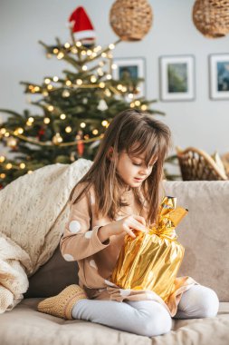 Kanepede oturan küçük bir kız elinde altın bir fiyonkla süslenmiş güzel bir altın hediye tutuyor. Arkasında, Noel Baba şapkasıyla süslenmiş ışıldayan bir Noel ağacı odaya sıcaklık ve şenlik katıyor.