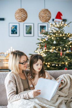 Bir anne ve kızı, Noel ağacının önünde açık bir kitapla oturup sıcak, aile odaklı bir an yaratıyorlar. Noel neşesi ve beraberliği ile dolu.