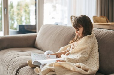 Yumuşak, krem renkli bir battaniyeyle kaplı genç bir kız kanepeye oturup kitap okuyor. Odanın sıcak atmosferi rahatlama duygusunu arttırıyor ve kitabına odaklanıyor..