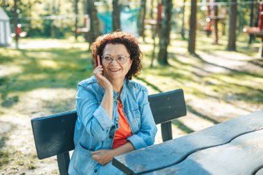 60 'lı yaşlarında gülümseyen bir kadın parktaki bir bankta rahatça oturup telefonuyla konuşuyor. Yeşil ağaçlı kot ceket giyiyor ve olay yerini çevreleyen sakin bir park ortamı var..