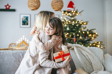 The living room is filled with holiday warmth as a woman and her daughter sitting on a couch and hugging, exchanging a gift, with a beautifully lit Christmas tree in the background clipart