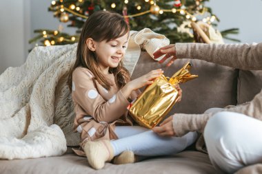 A cute little girl receiving a Christmas present from her mother while they sit together on a couch. The room lit by a Christmas tree adorned with lights and ornaments clipart