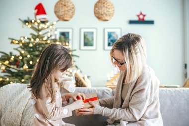 A woman and her daughter are seated on a couch, a girl is opening a holiday gift, as a Christmas tree adorned with ornaments and lights glows warmly in the background clipart