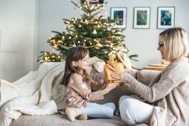 Seated on a cozy couch, a woman hands a gift to her little daughter in front of a festive Christmas tree, surrounded by holiday decorations, filling the room with warmth and joy clipart