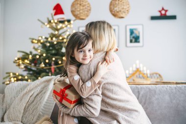 A mother and her little daughter hugging while exchanging a present on a couch, the Christmas tree behind them glowing with holiday lights clipart