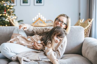 Bir kadın ve kızı kanepede sıcak bir kucaklaşmayı paylaşıyor, arka planda süslü bir Noel ağacının parıltısı altında gülüyorlar, neşe ve aile bağlarıyla dolu sıcak bir tatil anını yakalıyorlar.