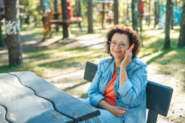 Parktaki bankta dinlenen, cep telefonuyla konuşan, mavi kot ceket ve turuncu bluz giyen olgun bir kadın. Arka planda huzurlu yeşil ağaçlar var..