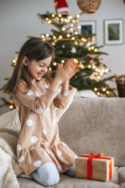 Çocuk, şenlikli bir oturma odasındaki koltukta diz çökmüş ellerini çırpıyor kırmızı kurdeleli bir hediyeye bakıyor. Arkaplanda ışık ve süslerle süslenmiş bir Noel ağacı yer alıyor.