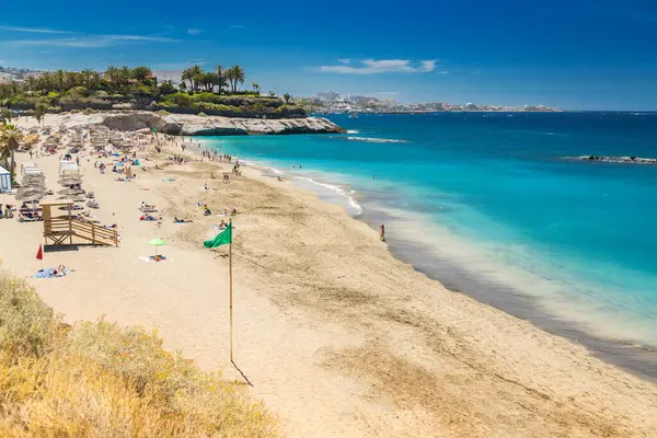 Playa del Duque, Tenerife 'de altın kum ve berrak sularla güneşli bir manzara. Yüzücüler ve güneş banyosu yapanlar sahili yeşil bir bayrak ile doldurdu..