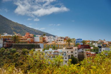 Tenerife 'deki resimli tepe kenti, düz çatıları olan renkli binalar. Ön planda yemyeşil ve arka planda bir dağ berrak, parçalı bulutlu bir gökyüzünün altında..