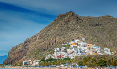 Tenerife 'deki San Andres köyünün, engebeli bir dağın eteklerine kurulmuş manzarası. Tepenin yamacında renkli evler kümelenir, ön planda palmiye ağaçları olan bir plaj vardır..