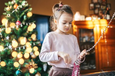 Süslü bir Noel ağacının önünde elinde süs ya da çelenk ile duran küçük bir kız. Noel ağacı çeşitli süsler ve ışıklarla süslenerek şenlikli bir atmosfer yaratır.