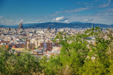 Şehrin göbeğinde Sagrada Familia ile Barselona 'nın panoramik bir görüntüsü, çeşitli binalar, yeşil alanlar ve uzak dağlarla çevrili.
