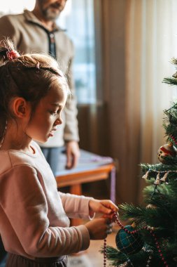 Küçük bir kız noel ağacını boncuklarla süslüyor. Dedesi arka planda durmuş, çocuğu izliyor..