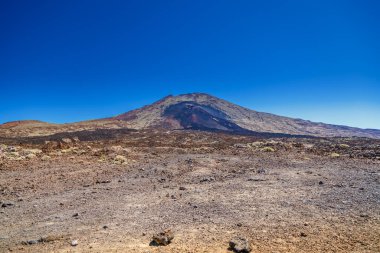 Açık mavi gökyüzünün altında geniş bir volkanik dağ olan çorak, kayalık bir arazi, kahverengi ve siyah volkanik kayaların çeşitli tonlarına sahip, Teide, Tenerife