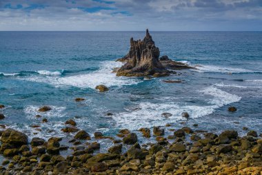 Tenerife, Playa Benijo 'daki sahil sahnesinde denizden yükselen belirgin kaya oluşumuyla, dalgalar ona çarpıyor, derin mavi ve turkuaz su, ve parçalı bulutlu gökyüzü
