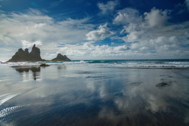 Playa Benijo 'da huzurlu bir sahil manzarası aynaya benzeyen bir okyanusu gösteriyor ve parçalı bulutlu bir gökyüzünün altında kayalıklara çarpıyor.