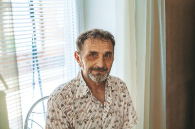 60-70 yaşlarında sakallı bir adam, desenli bir gömlek giymiş, pencerenin yanında oturmuş, doğal ışığın içeri girmesine izin veren perdeler, arka planda açık renkli perdeler.