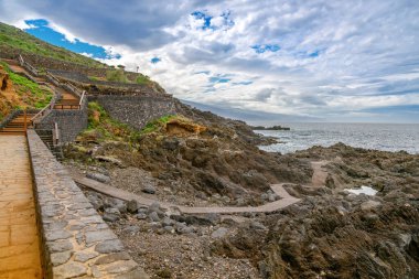 El Sauzal, Tenerife 'deki engebeli kıyı şeridi, taştan yollar ve teraslı yamaçlarda yemyeşil bitki örtüsü. Okyanus üzerindeki kasvetli gökyüzü manzaraya dramatik bir atmosfer ekler.