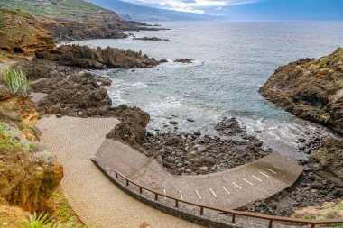 El Sauzal, Tenerife 'deki manzaralı kıyı manzarası kayalık kıyı şeridine giden dolambaçlı bir yol gösteriyor. Parlak bir gökyüzü altında engebeli kayalar ve sakin okyanus dalgalarıyla tezat oluşturan canlı yeşiller.