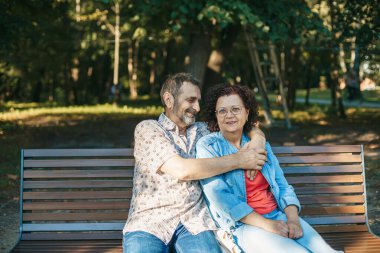 Sevecen yaşlı çift ağaçlarla çevrili yeşil bir parktaki ahşap bankta ve doğanın huzuruyla birlikte kaliteli zaman geçiriyorlar.