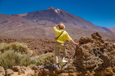 Dişi bir gezgin, sarı gömleği ve hasır şapkası engebeli volkanik manzaraya karşı durarak Teide Dağı 'nın hayranlık uyandıran manzarasını izlemek için durur.