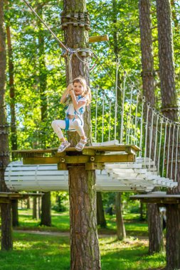Koşumlu küçük bir çocuk macera parkında bir halat pistinde geziniyor. Çocuk heyecan ve güven gösterirken, yemyeşil bir orman ormanıyla çevrili.