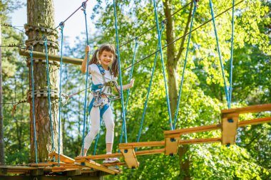 Bir macera parkında ipler üzerinde gezinen genç bir kız güvenli bir emniyet kemeri takmış ve yemyeşil ağaçlarla çevrili platformlar arasında dengeli bir şekilde gülümsüyor.