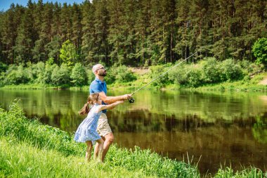 Baba, kızına yemyeşil sularla çevrili güneşli bir günde nasıl balık tutulacağını öğretiyor. Etkileşim, doğal ortamdaki bağ, öğrenme ve zevki yakalar..