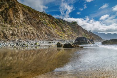 Sahildeki kayalıkların manzarası Playa Benijo, Tenerife 'deki sakin denizi yansıtıyor. Kayalık kıyı şeridi ve sakin sular canlı mavi gökyüzünün altında sakin ve resimsel bir manzara yaratır.