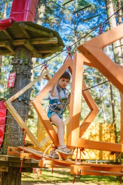 Macera parkında zorlu bir halat kursunda gezinen ve emniyet için koşum takımı takan küçük bir çocuk. Ağaçlarla çevrili ahşap yapılar üzerinde dengelenirken cesaret ve konsantrasyonu gösterir