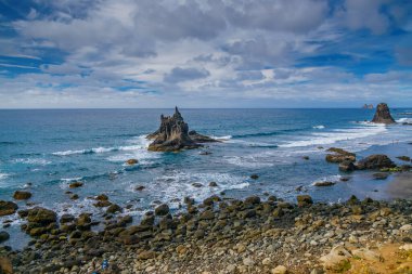 Playa Benijo, Tenerife 'nin çakıllı kıyı şeridi boyunca büyük kaya oluşumlarının yer aldığı kıyı sahnesi. Dalgalar parçalı bulutlu bir gökyüzünün altında kayalara çarpar ve dramatik bir deniz manzarası yaratır.