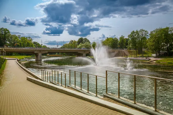 Litvanya, Anyksciai 'deki Riverside sahnesinde sakin bir nehrin üzerinde çeşmeli bir köprü sergileniyor. Asfaltlı bir yürüyüş yolu yan yana uzanır, bulutların altında sakin yürüyüşler yapmaya davet eder.