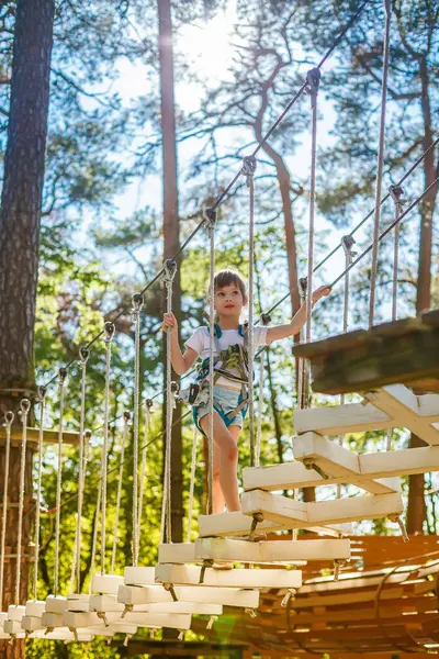 Genç bir çocuk macera parkında güven içinde ip köprüden geçerken cesaret ve denge gösterisi yapıyor. Güneş ışığı ağaçların arasından süzülüp, canlı bir açık hava manzarası yaratıyor.