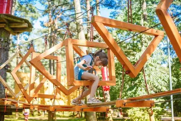 Genç kız, macera parkındaki bir halat kursunda güven içinde dengede duruyor. Emniyet kemeri takarak, yemyeşil ağaçlar ve yemyeşil ağaçlarla çevrili ahşap geometrik çerçevelerde geziniyor.