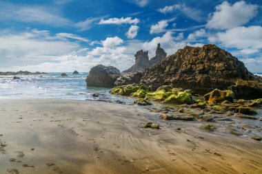 Tenerife 'deki Playa Benijo kumsalındaki ayak izleri yeşil yosunlarla kayalık oluşumlara yol açıyor, açık mavi gökyüzünün altında dağınık bulutlar, manzaralı bir kıyı manzarası tasvir ediyor.