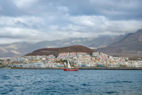 Los Cristianos kıyı kasabası ile mavi suları geçen kırmızı yelkenli dramatik bulutlu gökyüzü ve engebeli dağ zemininin altında yamaçlara yayılıyor.