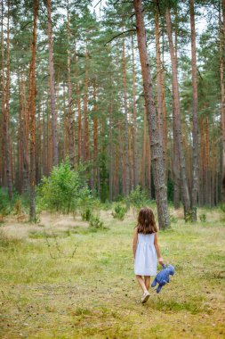 Küçük bir çocuk yoğun bir çam ormanında yürüyor, açık mavi bir elbise giyiyor ve elinde mavi bir oyuncak ayı tutuyor.
